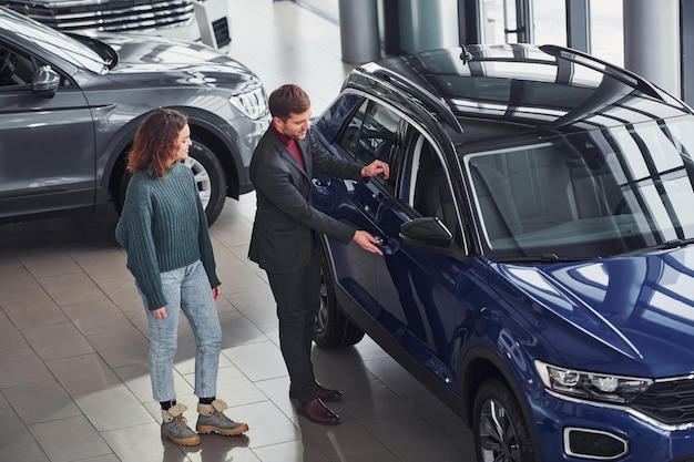 Professional salesman assisting young girl by choosing new modern automobile indoors.