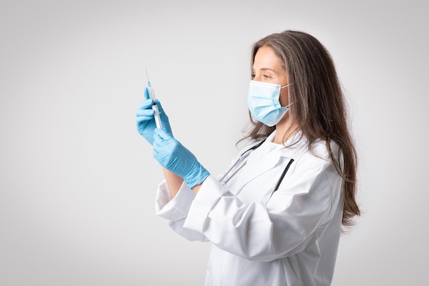 Professional senior female doctor in coat protective mask gloves with syringe and vaccine posing on