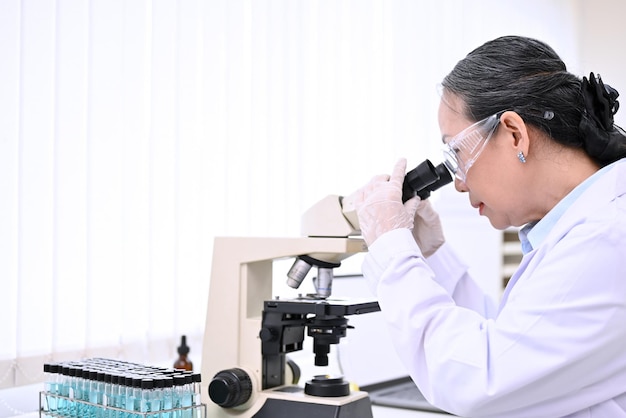 Professional senior female scientist looking under Microscope analyzing samples in the lab