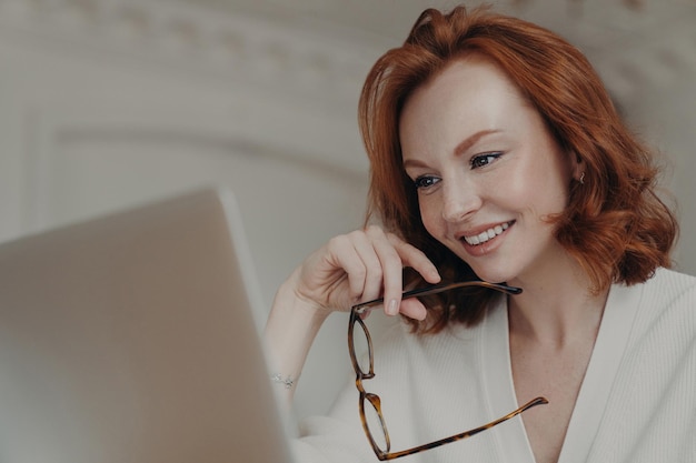 Professional smiling ginger female web designer uses application on laptop computer for creating project work makes online research and browses internet holds spectacles has happy expression