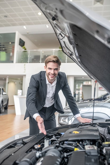 Professional. smiling young adult affable man in white shirt\
and dark suit showing with hand in open hood of car in\
dealership