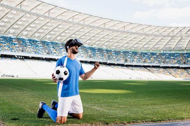Professional soccer player in vr headset and blue and white uniform with ball standing on knees and