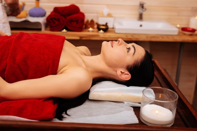 Professional SPA salon. Peaceful dark-haired woman covered in red towel resting on wooden table in massage cabinet