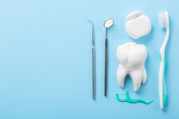 professional steel dental instruments with a mirror near white tooth model, toothbrush and dental floss on light blue table.