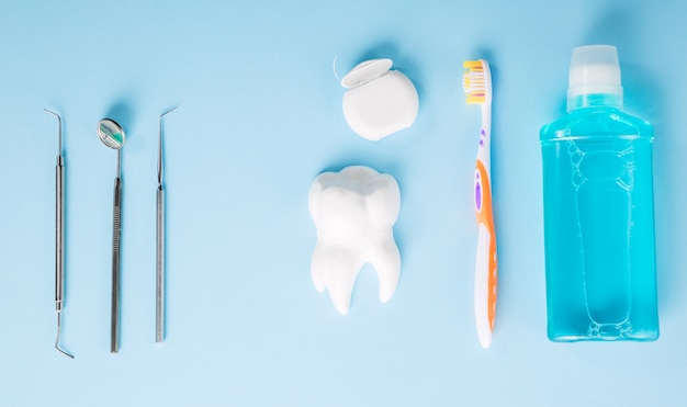 Professional steel dental instruments with a mirror near white tooth model, toothbrush, dental floss and mouthwash on light blue table.