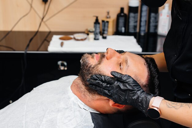 현대적인 세련된 이발소의 전문 스타일리스트가 젊은 남자의 머리카락을 면도하고 자릅니다.