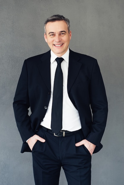 Professional and successful. Mature businessman keeping hands in pockets and looking at camera with smile while standing against grey background