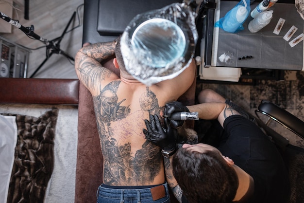 Il maestro del tatuaggio professionista con una macchinetta per tatuaggi riempie il disegno di un cavaliere sulla schiena di un uomo, vista dall'alto. tatuatori del flusso di lavoro