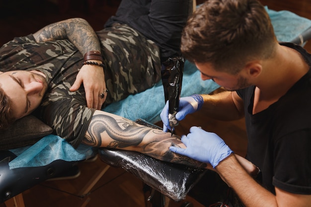 Профессиональный татуировщик делает татуировку на руке молодого человека на машине черными чернилами
