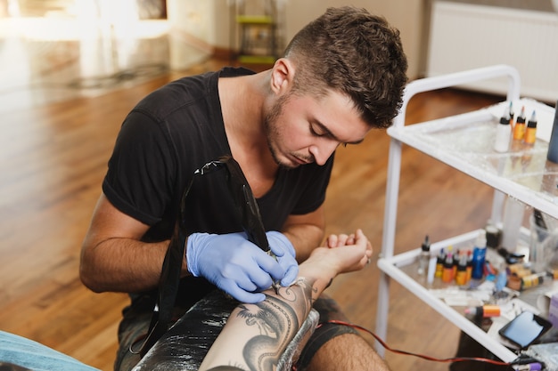 A professional tattooer artist doing tattoo on the arm of a young man by machine with black ink