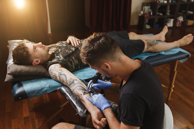 A professional tattooer artist doing tattoo on the arm of a young man by machine with black ink