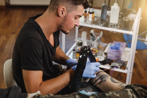 A professional tattooer artist doing tattoo on the arm of a young man by machine with black ink