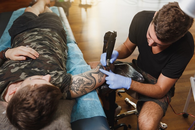 A professional tattooer artist doing tattoo on the arm of a young man by machine with black ink