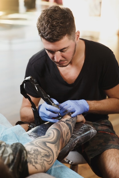 A professional tattooer artist doing tattoo on the arm of a young man by machine with black ink