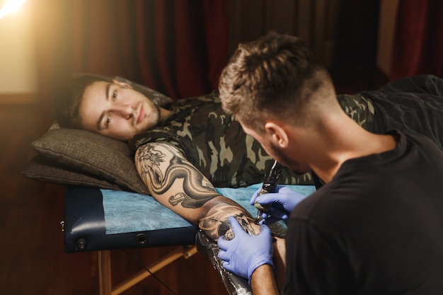 A professional tattooer artist doing tattoo on the arm of a young man by machine with black ink