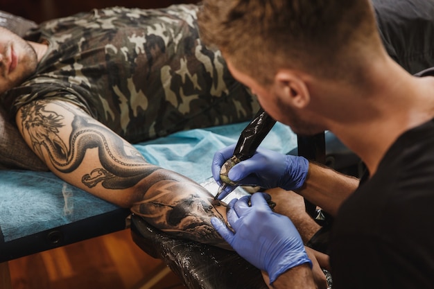 A professional tattooer artist doing tattoo on the arm of a young man by machine with black ink