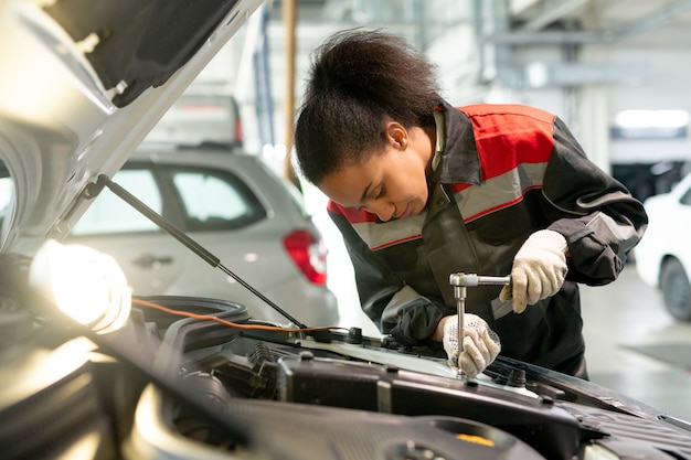 Professional technician of car maintenance service standing by open engine compartment in workshop and using handtool to fix small details