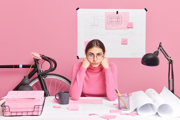 Professional tired female architect works on building sketch\
feels tired of long hours working checks graphic outlay tries to\
improve idea of planning poses at desktop surrounded with papers\
stickers