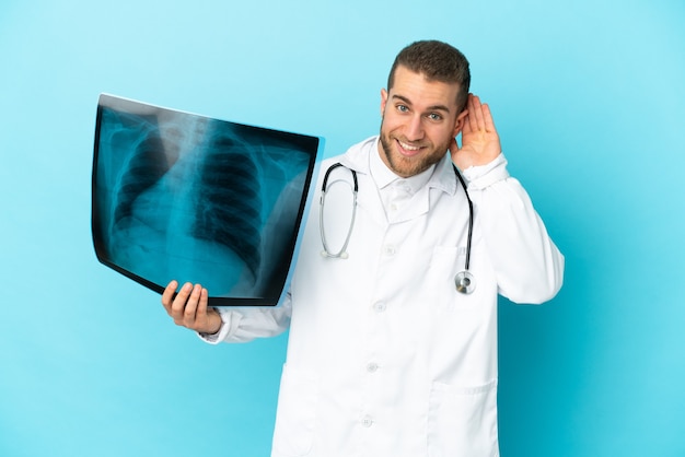 Professional traumatologist isolated on blue listening to something by putting hand on the ear