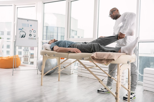 Professional treatment. Skillful nice doctor holding his patients leg while doing rehabilitation therapy