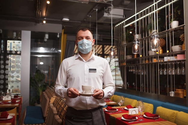 Professional waiter working at the restaurant during coronavirus pandemic, wearing medical face mask