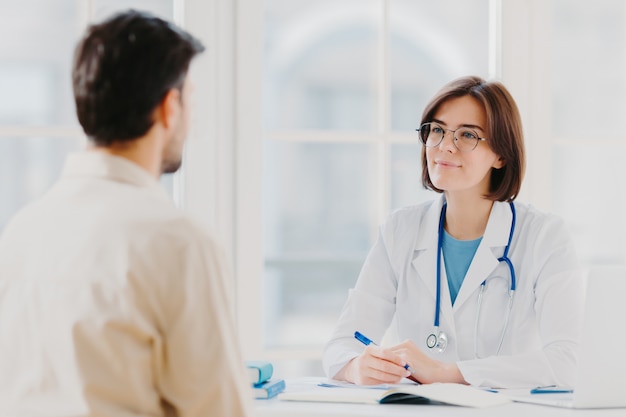 Professional woman doctor speaks with male patient, gives consultancy and makes prescription