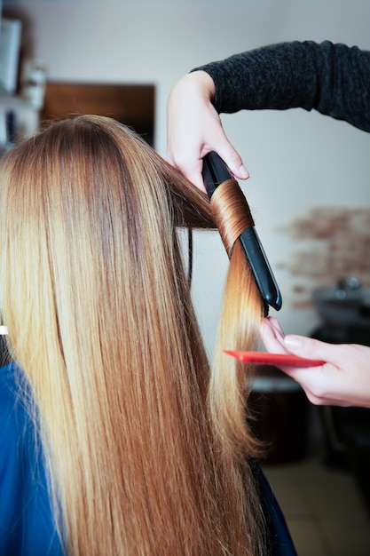 professional woman hairdresser making hairstyle using curling iron for long hair of young female in beauty salon.