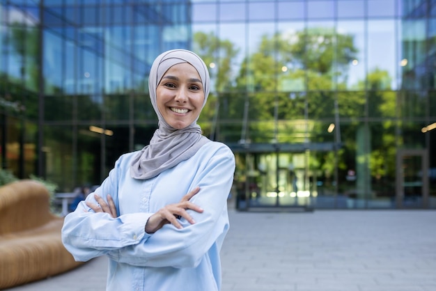 A professional woman in a hijab stands confidently with crossed arms outside a contemporary office