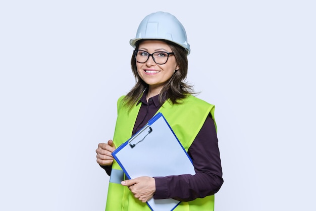 Professional woman in safety vest helmet holding clipboard on white background