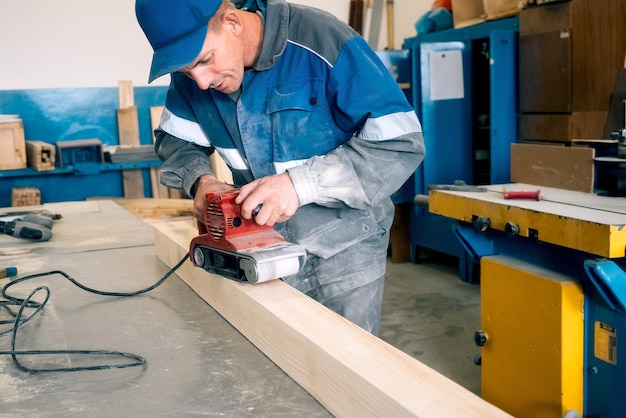 Professional woodworker grinds wooden bar on workbench in carpentry workshop with tool Middleaged worker in overalls processes wood with electric grinder Real workflow