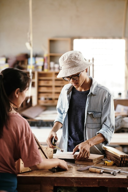 ワークベンチでプロジェクトに取り組んでいるときに巻尺で木の板を測定するプロの若いアジアの大工