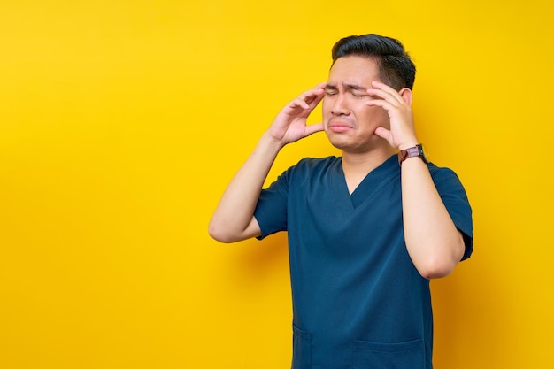 Professional young asian male doctor or nurse wearing a blue uniform feeling sick and suffering from headache or migraine isolated on yellow background healthcare medicine concept
