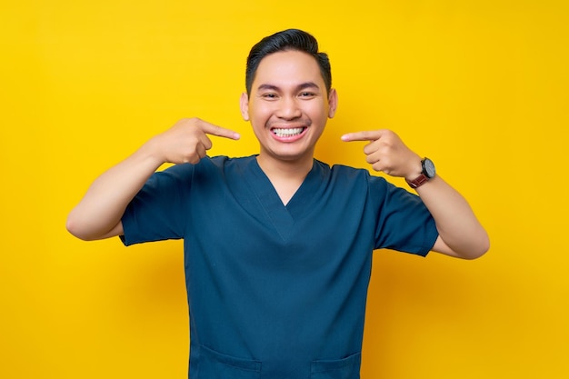 Professional young asian male doctor or nurse wearing a blue uniform showing wellgroomed white teeth isolated on yellow background healthcare medicine concept