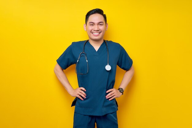 Professional young asian male doctor or nurse wearing a blue uniform standing confident with hand on waist and smiling friendly isolated on yellow background healthcare medicine concept