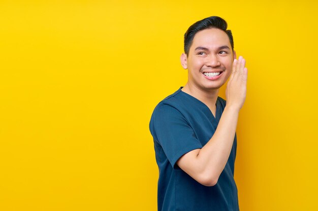 Professional young asian male doctor or nurse wearing a blue uniform whispering gossip in the hospital isolated on yellow background healthcare medicine concept