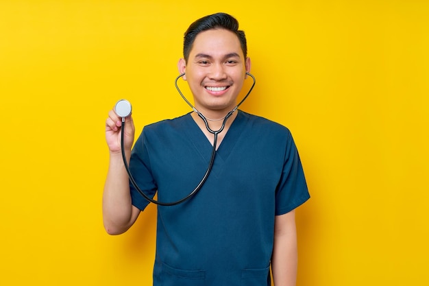 Photo professional young asian man doctor or nurse wearing a blue uniform demonstrates using stethoscope isolated on yellow background healthcare medicine concept