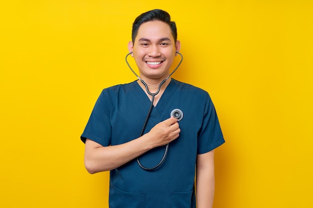 Professional young asian man doctor or nurse wearing a blue uniform listening to lungs with a stethoscope doing medical checkups in a clinic isolated on yellow background healthcare medicine concept
