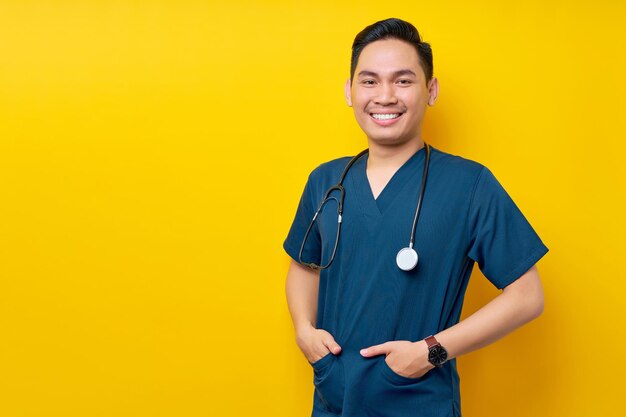 Photo professional young asian man doctor or nurse wearing a blue uniform and stethoscope standing confidently while smiling friendly at camera hands in pockets isolated on yellow background