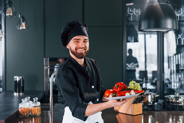 Professional young chef cook in uniform holds plate with vegetables on the kitchen