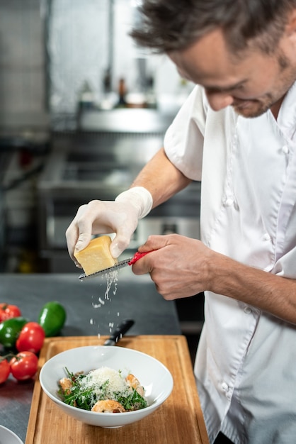 Молодой профессиональный шеф-повар натирает свежий сыр в керамическую миску с салатом, состоящим из рукколы, вареных креветок и других ингредиентов.