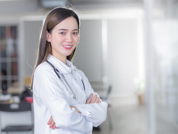病院の診察室で笑顔で腕を組んで立っているプロの若い女性医師