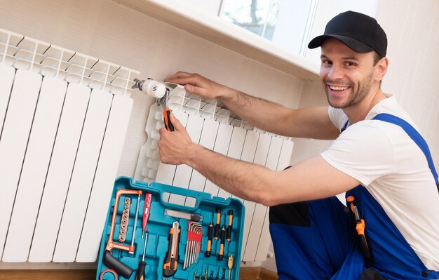 Professional young repairman in special uniform with tools is installing radiators and thermostat in domestic room