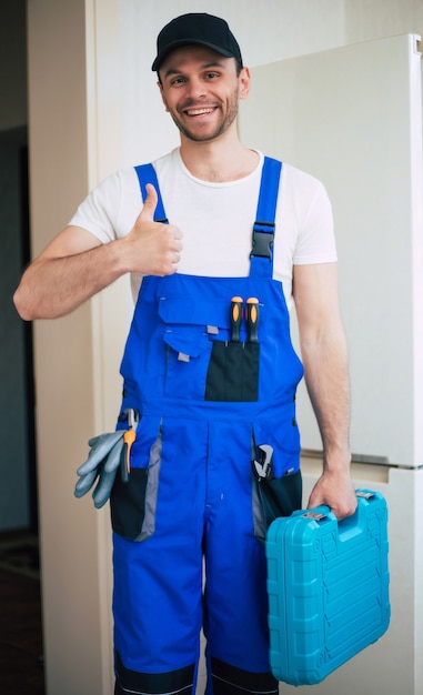 Профессиональный молодой ремонтник в рабочей форме и кепке с современным ящиком для инструментов с оборудованием после ремонта холодильника на кухне