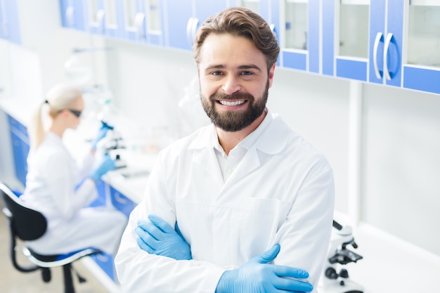 Professioneel lab. Slimme zelfverzekerde mannelijke wetenschapper permanent cross overhandigd en glimlachen tijdens het werken in het lab