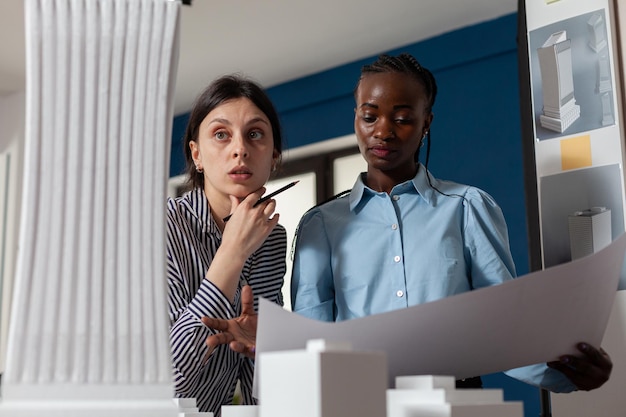 Professioneel team van twee architecten die het wolkenkrabbermodel van wit schuim inspecteren terwijl ze blauwdrukken vasthouden. Collega's van projectingenieurs die geïnspireerd kijken voor 3d residentiële maquette.