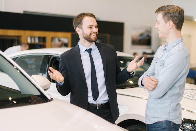 Foto professionele autoverkoper die een pak draagt, vertelt geïnteresseerde kopers over de auto in de autoshowroom. concept van het kiezen en kopen van een nieuwe auto in de showroom.