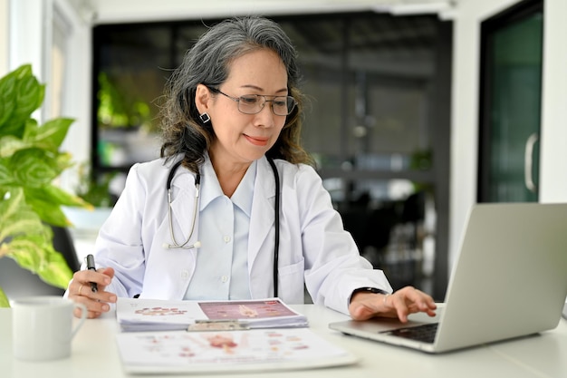 Professionele Aziatische oude vrouwelijke arts die in het kantoor van de dokter werkt met behulp van laptop