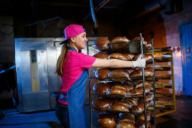 Professionele bakker - een jonge, mooie vrouw in een jeansschort houdt vers brood vast tegen de achtergrond van een bakkerij of bakkerij. bakkerijproducten. broodproductie