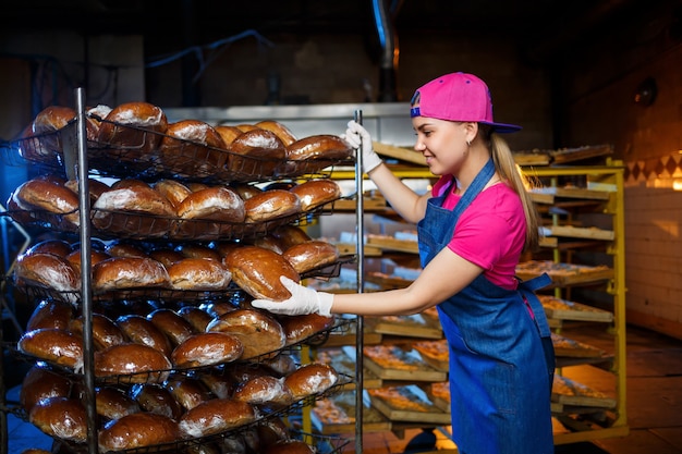 Professionele bakker - een jonge, mooie vrouw in een jeansschort houdt vers brood vast tegen de achtergrond van een bakkerij of bakkerij. Bakkerijproducten. Broodproductie