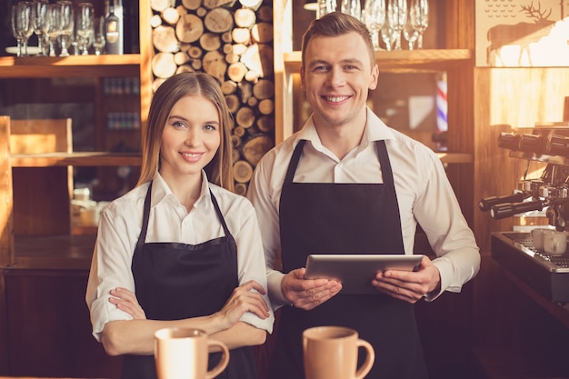 Professionele barista. Jonge vrouw en man in schorten kijken naar de camera, glimlachen en staan aan de toog. Ze gebruiken een tabletcomputer